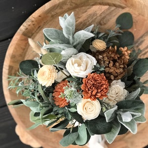 May include: A rustic wedding bouquet with cream, orange, and brown flowers, greenery, and dried elements.