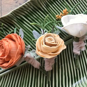 May include: Three wooden flower boutonnieres in peach, cream, and white. The flowers are tied with a light pink ribbon and have green foliage.