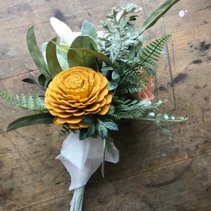 May include: A bouquet of faux flowers with a yellow wooden flower, green ferns, and white and gray foliage. The bouquet is wrapped in white fabric and tied with a ribbon.