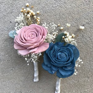 May include: Two boutonnieres with wooden rose flowers. One is dusty rose, the other is blue. Both have white baby's breath and are wrapped with white ribbon. Perfect for a wedding or special event.