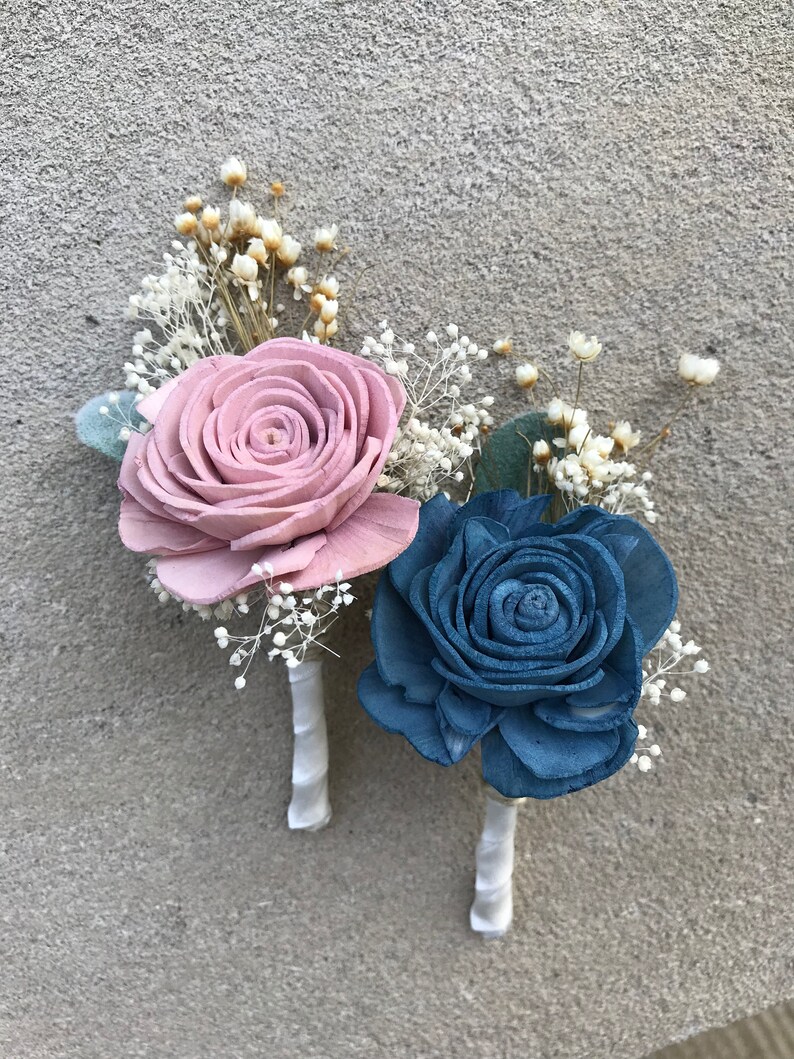 May include: Two boutonnieres with wooden rose flowers. One is dusty rose, the other is blue. Both have white baby's breath and are wrapped with white ribbon. Perfect for a wedding or special event.