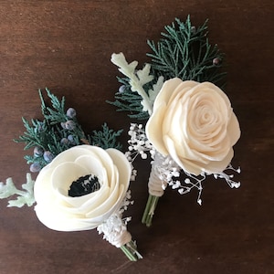 May include: Two white sola wood flower boutonnieres with greenery and white baby's breath.