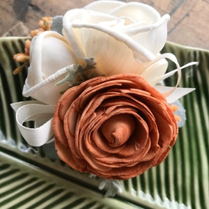 May include: A bouquet of three wooden roses, one orange and two white, with green leaves and a white ribbon. The roses are arranged on a green and white striped surface.
