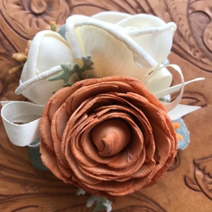 May include: A brown wooden rose with white fabric roses and greenery. The roses are arranged in a bouquet and tied with a white ribbon.