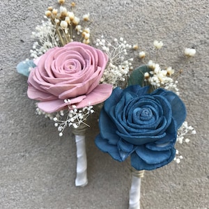 May include: Two boutonnieres with wooden rose flowers. One is a dusty rose color, the other is a muted blue. Both have white baby's breath and greenery accents. The stems are wrapped in twine and white ribbon.