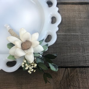 May include: A white flower boutonniere with greenery and small white flowers. The flower is made of wood and has a natural brown center.