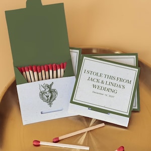 May include: Olive green matchbox with red-tipped matches, featuring a vintage cherub illustration. The matchbox is open, revealing the matches, and is next to a card that reads "I STOLE THIS FROM JACK & LINDA'S WEDDING December 12, 2027."