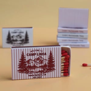 May include: A rectangular matchbox with a maroon and white striped pattern, featuring a graphic of mountains and pine trees. The text reads Welcome to Camp Linda, Santa Barbara, CA, Est. 2027. The box is partially open, revealing wooden matches with red tips.