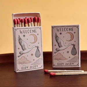 May include: Two rectangular matchboxes featuring a white background with a black line drawing of a stork carrying a bundle, a crescent moon, stars, and clouds. The text reads "WELCOME" and "BABY AVERY". One box is open, revealing red-tipped matches.