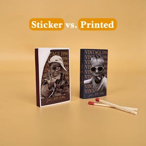 May include: Two rectangular matchboxes with custom photo designs labeled Sticker vs. Printed. One box features a peeling sticker showing a person in sunglasses, while the other has a direct print. Two wooden matches with red tips lie nearby.