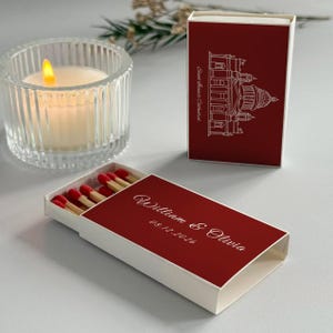 May include: A lit candle in a clear glass holder and two matchboxes. One matchbox is open, revealing red-tipped matches. The matchboxes are red with white text and a cathedral illustration. The text reads "William & Olivia" and "08.12.2026".