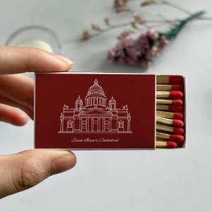 May include: A red matchbox featuring a white line drawing of Saint Isaac's Cathedral. The text "Saint Isaac's Cathedral" is below the illustration. The matchbox is open, revealing several matches with red tips.