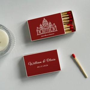 May include: Two red matchboxes with white text and illustrations. One box features a cathedral drawing and the text "Saint Isaac's Cathedral." The other box has the names "William & Olivia" and the date "08.12.2026." A single match is next to the boxes.