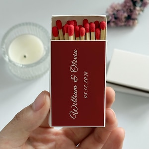 May include: A matchbox with a deep red cover, featuring the names "William & Olivia" and the date "08.12.2026" in elegant white script. The box is open, revealing red-tipped matches.