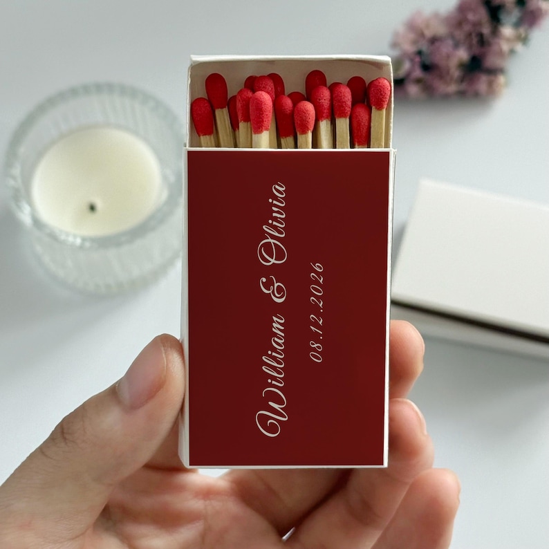 May include: A matchbox with a deep red cover, featuring the names "William & Olivia" and the date "08.12.2026" in elegant white script. The box is open, revealing red-tipped matches.