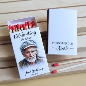 May include: Two white matchboxes with red-tipped matches. One box is open, displaying a portrait of an older man in a cap with the text Celebrating the life of Jack Anderson 1938-2027. The closed box features the text FOREVER IN OUR Heart.