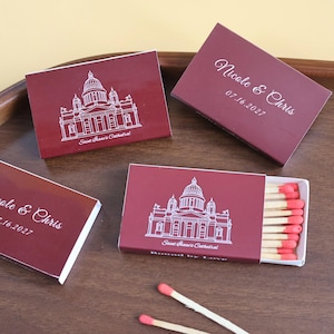May include: Four rectangular matchboxes in deep burgundy with white line art of Saint Isaac's Cathedral. Two boxes feature the text Nicole & Chris 07.16.2027. One open box reveals wooden matches with pink tips. Two loose matches lie on the surface.