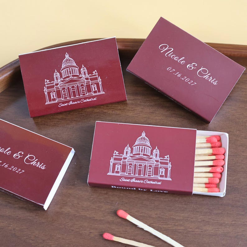 May include: Four rectangular matchboxes in deep burgundy with white line art of Saint Isaac's Cathedral. Two boxes feature the text Nicole & Chris 07.16.2027. One open box reveals wooden matches with pink tips. Two loose matches lie on the surface.