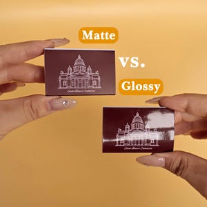 May include: Two rectangular matchboxes with a dark maroon finish, one matte and one glossy. Both feature a white line-art illustration of Saint Isaac's Cathedral and the text "Saint Isaac's Cathedral". The words "Matte vs. Glossy" appear in yellow.