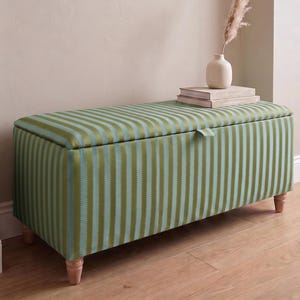 May include: A rectangular storage ottoman upholstered in a fabric featuring vertical stripes of olive green and light blue. The piece has a hinged lid with a small fabric pull tab and is supported by four short, light-coloured wooden legs.