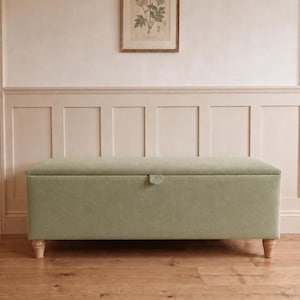 May include: A rectangular storage bench upholstered in a solid sage green fabric. The bench features a hinged lid with a small fabric pull tab centred on the front edge and is supported by four short, light-coloured wooden legs with a fluted design.