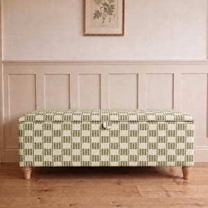 May include: A rectangular storage bench upholstered in a cream and olive green checkered fabric with a small fabric pull tab on the front lid. The bench features four short, light-toned wooden legs and a structured, boxy silhouette.