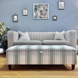 May include: A rectangular storage ottoman featuring a striped pattern in shades of sage green, light blue, and cream. The ottoman has a hinged lid with a small fabric pull tab and is supported by four dark brown tapered wooden legs.