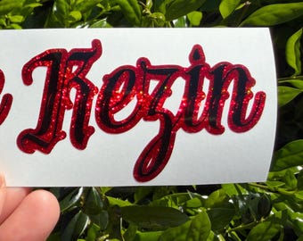 May include: A rectangular white decal featuring the text Unique Kezin in a stylized, bold script font. The lettering has a deep red glitter finish with a thick black outline, creating a high-contrast appearance against the white background.