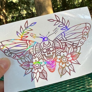 May include: A holographic sticker featuring a detailed line drawing of a butterfly with a skull in the center. The butterfly has intricate floral designs on its wings and a crescent moon above the skull.