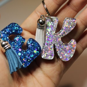 May include: Two glittery resin keychains in the shape of the letters "E" and "K". The "E" is blue with a matching tassel, and the "K" is silver with a white tassel. Both keychains have silver key rings.