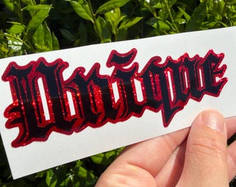 May include: A rectangular white adhesive decal featuring the text Unique Rezin in a bold, black, gothic-style font with a shimmering red glitter outline. The lettering is arranged horizontally on a smooth, flat surface.