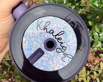 May include: A purple tumbler with a black lid featuring a circular insert with a light blue, pink, and white speckled glitter pattern. The name Khaleigh is written in black cursive script across the center of the glittery insert.