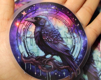 May include: A round sticker with a galaxy background and a black raven perched on a branch. The raven has iridescent purple and blue feathers. The sticker has a glossy finish.