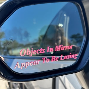 May include: A close-up of a car's side view mirror with a pink sticker that reads "Objects In Mirror Appear To Be Losing".