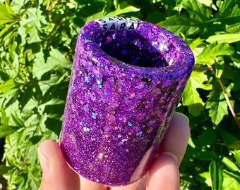May include: A cylindrical, purple glitter resin shot glass with the name "SARA" in white letters. The shot glass is held in a hand against a backdrop of green foliage.