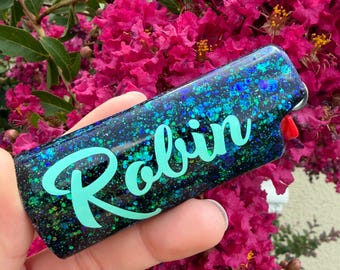 May include: A rectangular lighter featuring a dark base covered in iridescent blue and green glitter. The name Robin is written across the center in a light teal script font. A small red ignition button is visible on the side of the lighter.