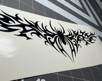 May include: A black vinyl decal featuring a symmetrical tribal spider design with elongated, pointed flame-like extensions stretching horizontally across a white backing sheet, positioned over a dark gray cutting mat with visible grid lines.