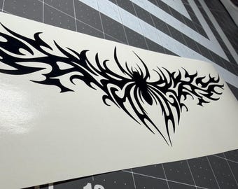 May include: A black vinyl decal featuring a symmetrical tribal spider design with elongated, pointed flame-like extensions stretching horizontally across a white backing sheet, positioned over a dark gray cutting mat with visible grid lines.