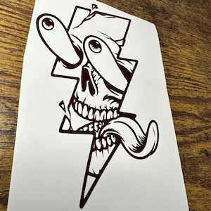 May include: A maroon colored sticker with a skull design. The skull is inside of a lightning bolt shape. The skull has one eye that is covered by a band and the other eye is bulging out. The skull is sticking its tongue out.