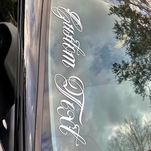 May include: A black car windshield with a white decal that reads "Custom Text" in an elegant script. The decal is centered and vertically oriented. The sky and trees are reflected in the glass.