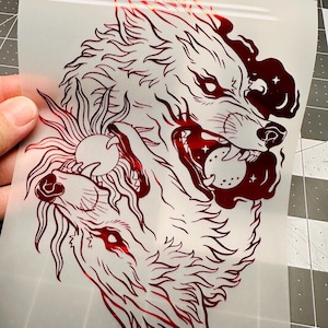 May include: A red wolf graphic with intricate details, featuring two wolf heads and a sun design. The artwork is on a transparent sheet, showcasing a detailed illustration suitable for various applications.