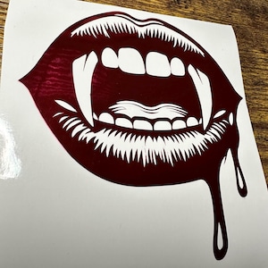 May include: A dark red vinyl decal featuring a graphic illustration of an open mouth with sharp vampire fangs and a single drop of liquid dripping from the lower lip. The design uses negative space to define the teeth and the tongue inside.