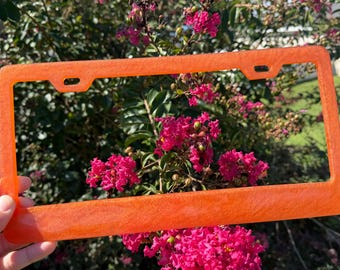 May include: A rectangular license plate frame with a vibrant orange, glittery finish. The frame features a smooth, glossy texture and includes two mounting holes at the top for attachment to a vehicle, showcasing a bright and reflective appearance.