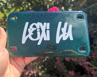 May include: A rectangular license plate frame with a dark teal and blue glitter finish. The surface features the white text LEXI LU in a stylized font. The frame includes four mounting holes, one in each corner, for attachment to a vehicle.