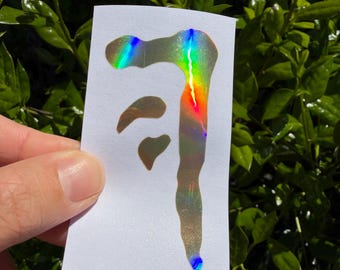 May include: A rectangular white sticker featuring a holographic, iridescent design that resembles a stylized, elongated abstract shape with a vertical drip-like element and two smaller, separate curved segments above it.