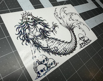 May include: A holographic sticker featuring a skeletal mermaid wearing a crown and shell top, surrounded by multiple skulls. The design is rendered in black lines with a shimmering, multi-colored iridescent finish on a white backing sheet.