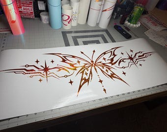 May include: A large, white adhesive decal sheet featuring a central butterfly design with intricate, sharp-edged wings. The butterfly and surrounding geometric starburst patterns are rendered in a vibrant, orange-red holographic glitter finish.