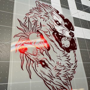May include: A red graphic of two wolves, one snarling with bared teeth, and the other with a sun-like design. The image is on a transparent background, suitable for heat transfer vinyl or similar applications.