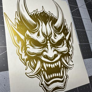 May include: A metallic gold vinyl decal featuring a detailed illustration of a Hannya mask with sharp horns, pointed ears, and bared teeth. The design is printed on a white backing sheet, showcasing intricate line work and a reflective finish.