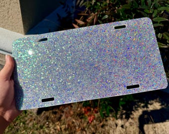 May include: A rectangular license plate frame featuring a dense, multi-colored holographic glitter finish. The surface reflects light with a spectrum of iridescent sparkles. It includes four pre-cut mounting slots positioned near the corners.
